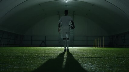 epic slow motion shot with walking footballer with ball in indoor arena, rear view in darkness