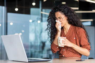 Sick woman working in office, hispanic woman having flu and cold drinking hot tea sneezing and coughing at workplace, using napkins, sitting at table with laptop.