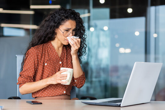 Sick woman working in office, hispanic woman having flu and cold drinking hot tea sneezing and coughing at workplace, using napkins, sitting at table with laptop.