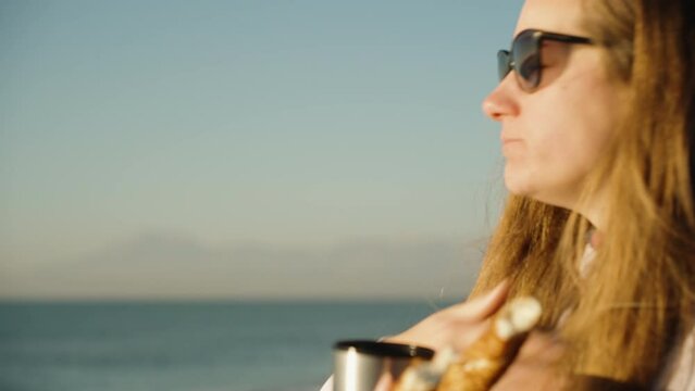 A girl on a cool morning on the seashore, Meets the sunset and eats a Turkish bagel Simit and drinks hot tea from a thermos. The wind blows her hair, she pulls it out of her mouth