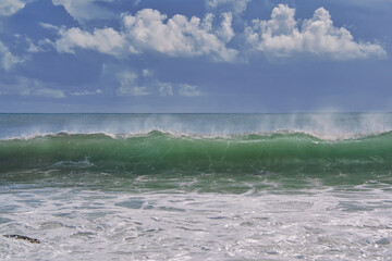 Turquoise wave breaking on a beach in sea with silver clouds
