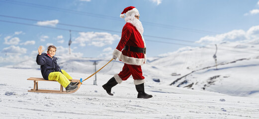 Full length profile shot of santa claus pulling a boy on a wooden sleigh at a mountain
