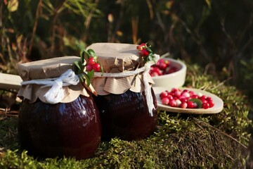 Fototapeta na wymiar Jars of delicious lingonberry jam and red berries outdoors, space for text