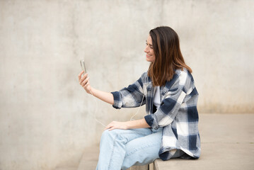 Girl black and white plaid shirt, jeans. Sitting on a bleacher. Making video call with headphones and cell phone.