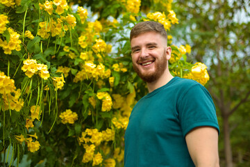 Portrait of happy handsome bearded guy, young positive man with beard is smelling beautiful yellow flowers in the garden, smiling, enjoying spring or summer day, breathing deep deeply fresh air