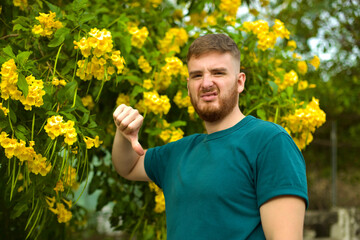 Naklejka na ściany i meble Portrait of handsome young allergic man is suffering from pollen allergy or cold on natural flowers, flowering tree background at spring or sunny summer day, sneezes, blowing his runny nose rubs eyes