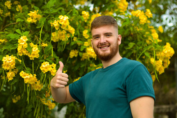 Portrait of happy handsome bearded guy, young positive man with beard is smelling beautiful yellow flowers in the garden, smiling, enjoying spring or summer day, breathing deep deeply fresh air