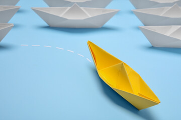 Yellow paper boat floating away from others on light blue background, closeup with space for text....