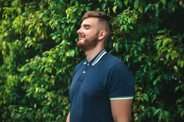 Portrait of a beautiful young bearded man with beard standing on an Ivy wall or in greenery,...