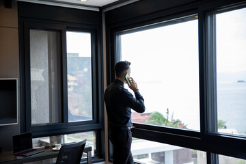 Portrait of Successful Businessman Wearing Suit Standing Using Smartphone Looking Out Window. Successful CEO planning e-commerce investment strategy.