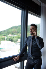 elegant man standing by window talking on cell phone, man wearing black suit answering his phone, copy space