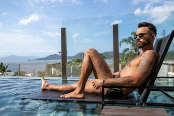 beautiful man inside the pool sitting on a chair, man with a big beard, swimming trunks and tattoos...