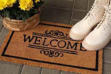 Door mat with word Welcome, stylish boots and beautiful flowers on floor
