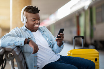 Happy Black Guy Using Smartphone While Relaxing oOn Bench At Railway Station