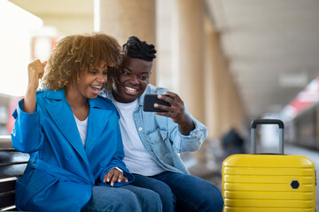 Online Win. Black Couple Celebrating Success With Smartphone At Railway Station