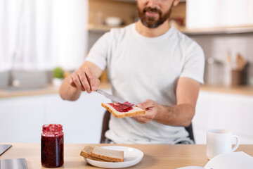 Smiling adult caucasian guy with beard making sandwich with jam at table, enjoy weekend in kitchen...