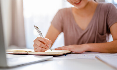 Smiling asian woman relaxing using technology of laptop computer while sitting on desk and writing into textbook, young creative girl working and typing on keyboard at home working at home concepts.