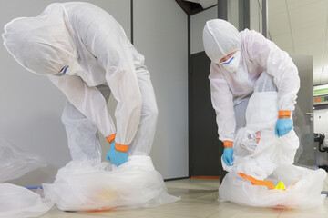 Operators in type 5/6 hazmat suits performing decontamination procedure, after asbestos incident, careful take off of protective suit in a disposal big bag