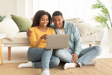 Happy millennial african american husband hug wife, watch video on laptop, have online call, sit on floor