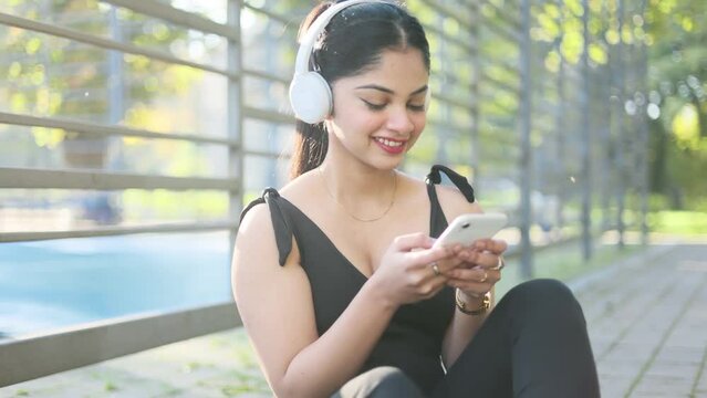 Close up beautiful indian woman hold smartphone, watching social media sitting near a stadium. Smiling young woman with headphones texting on her phone.	