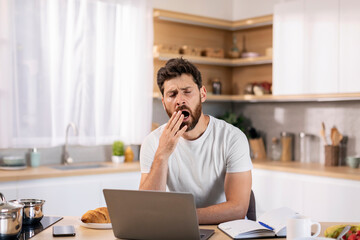 Sleepy tired bored handsome millennial caucasian guy with beard yawns, working at computer at table