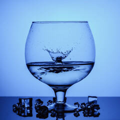 Splash drops of water in a glass goblet and ice on a blue background.