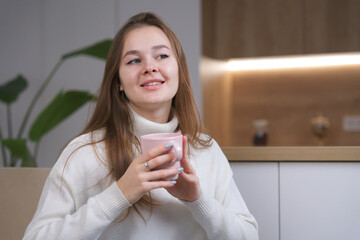 Young happy beautiful woman in sweater sitting in comfortable armchair at home in kitchen, smile drinking hot tea or coffee from cup