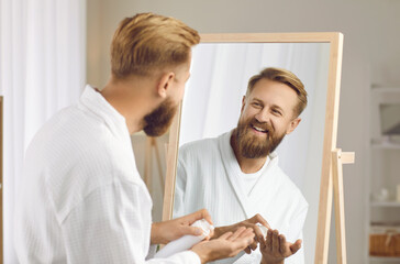 Grooming products for men. Happy bearded man with beard lotion in his hands enjoys morning routine of self care. Man in bathrobe looks at his reflection in the mirror. Morning male hygiene concept.