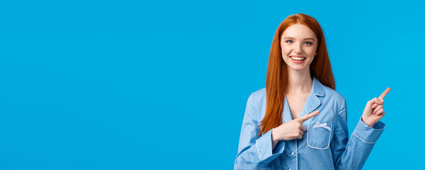 Fototapeta na wymiar Friendly attractive redhead female student showing person way in dormitory college, pointing right and smiling wearing nightwear, going sleep, showing her daily skincare routine, blue background