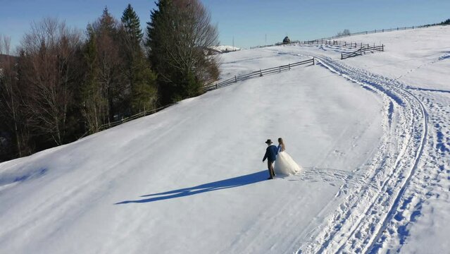 4k Top Aerial View Wedding Couple In Dress Denim Jacket Hat Walking Running On Snowy Carpathian Mountain. Loving Caucasian People Together At Winter In Ukraine. 4k Drone View From Above House Forest