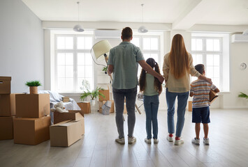 Fototapeta na wymiar Young family moving into new house or apartment. Mom, dad and children standing in spacious modern light living room full of cardboard boxes. Back view. Mortgage, buying new house, real estate concept