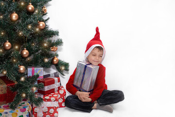 cute boy near the christmas tree holding a gift. happy child on a white background in a red New Year's hat and a red jacket hugs a gift