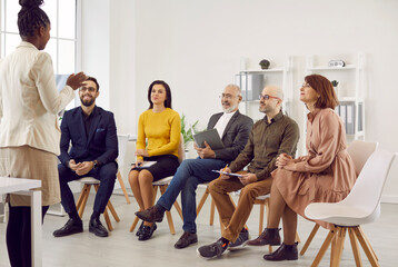 Female business coach speaks in front of small group of business people teaching them new business knowledge. Satisfied different people sitting on chairs in row and listening to dark skinned woman.