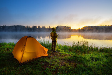 Fisherman on the bank of foggy river near an orange tent in the early morning