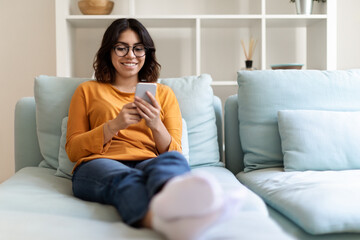 Beautiful Arab Female Wearing Eyeglasses Relaxing On Couch With Smartphone