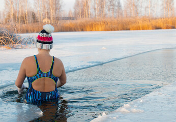 A woman in a swimsuit in a warm hat enters the hole in winter swimming, winter therapy, ice swimming in the river