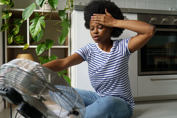 Unhappy African woman touching forehead having hot flashes using electric fan to cool off,...