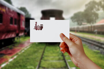 Man's hand holding train symbol paper on railway station. Concept of journey, travel, dream, freedom. Hand is holding paper train against rail road with empty space for text. Blurred background.