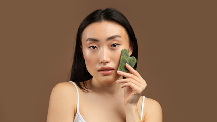Korean lady massaging face by gua sha quartz stone scraper for skincare, posing over brown...