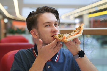 Portrait of young happy handsome man, hungry guy is sitting on food court in shopping mall, eating, holding in hands juicy tasty piece of pizza, biting. Fast junk unhealthy fat food. Hunger.