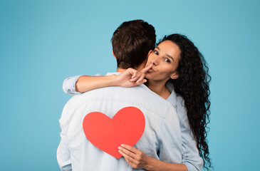 Cheerful young arab wife hugs european husband, holds heart, shows finger sign shhh on blue background