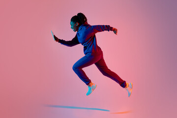 Fototapeta na wymiar Full body length shot of modern sporty lady jumping or running using cellphone over pink neon background, side view