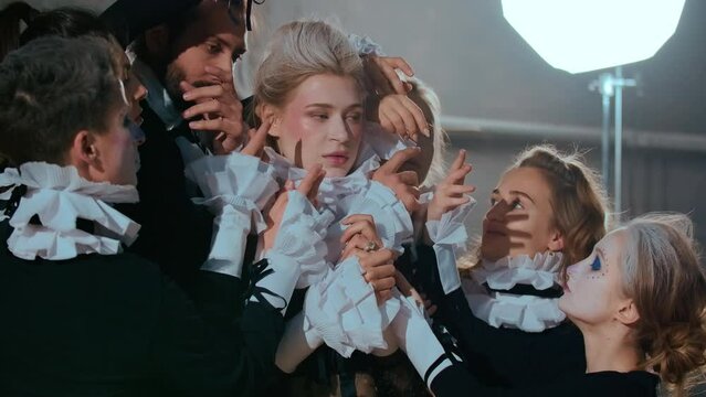 Portrait of impassive beautiful woman in theatrical costume looking away as group of actors stretching hands to lady. Professional Caucasian actress with expressionless facial expression rehearsing