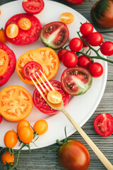 Delicious raw fresh colorful tomatoes of different shapes on white round plate with fork on wooden background. Chopped red and yellow tomatoes. Healthy food rich in vitamin C