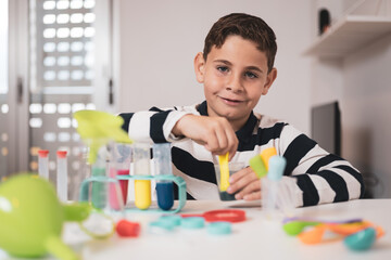 Boy having fun with chemistry lab in his living room. Science experiments at home for school homework. The little one mixes a liquid inside a test tube while looking at the camera.