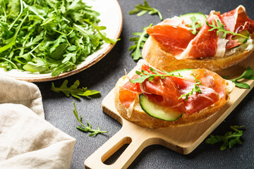 Open sandwiches with cream cheese, prosciutto, cucumber and arugula at cutting board.
