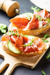 Appetizer crostini, open sandwiches with cream cheese, prosciutto, cucumber and arugula at cutting board.