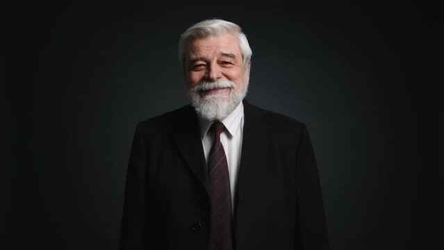 Close-up view of retired businessman wearing black formal suit. Portrait of happy, smiling man with gray beard looking at camera with black isolated background behind. High quality 4k footage