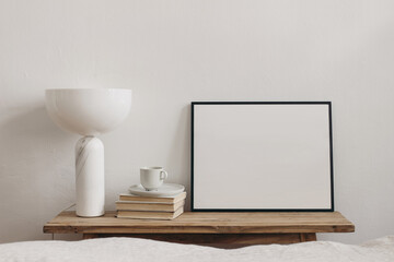 Modern Scandinavian bedroom. Minimal home, interior still life photo. Trendy marble lamp on old wooden bench, table. Blank black horizontal picture frame mockup. Cup of coffee on books. Blurred bed.