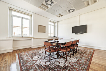 Vintage style office with rectangular wooden meeting table with wooden chairs upholstered in brown leather on a brown carpet and tv on the wall